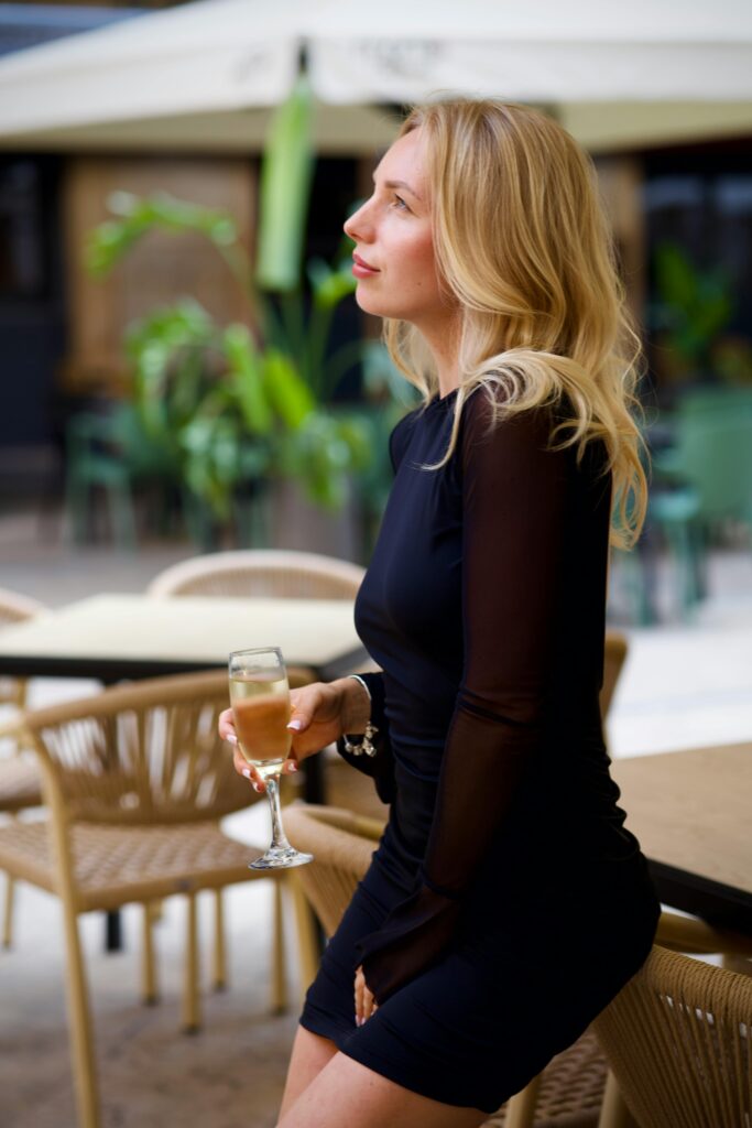 pexels-photo-34402742-34402742 Blonde woman in black dress drinking champagne at outdoor café